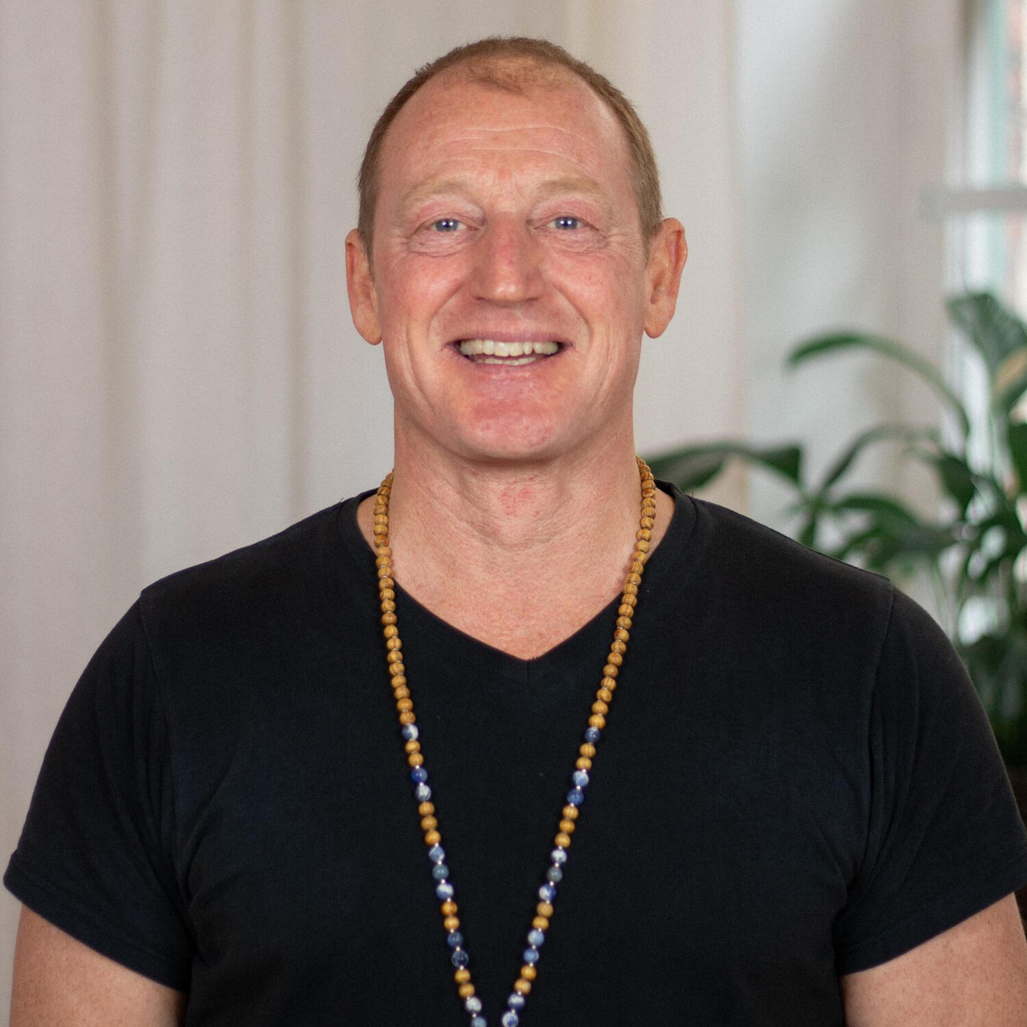 Yoga Kendra Malmö Vinyasa, Hatha, Yin Man with short hair, black t-shirt and wooden pearl necklace smiling into camera showing teeth