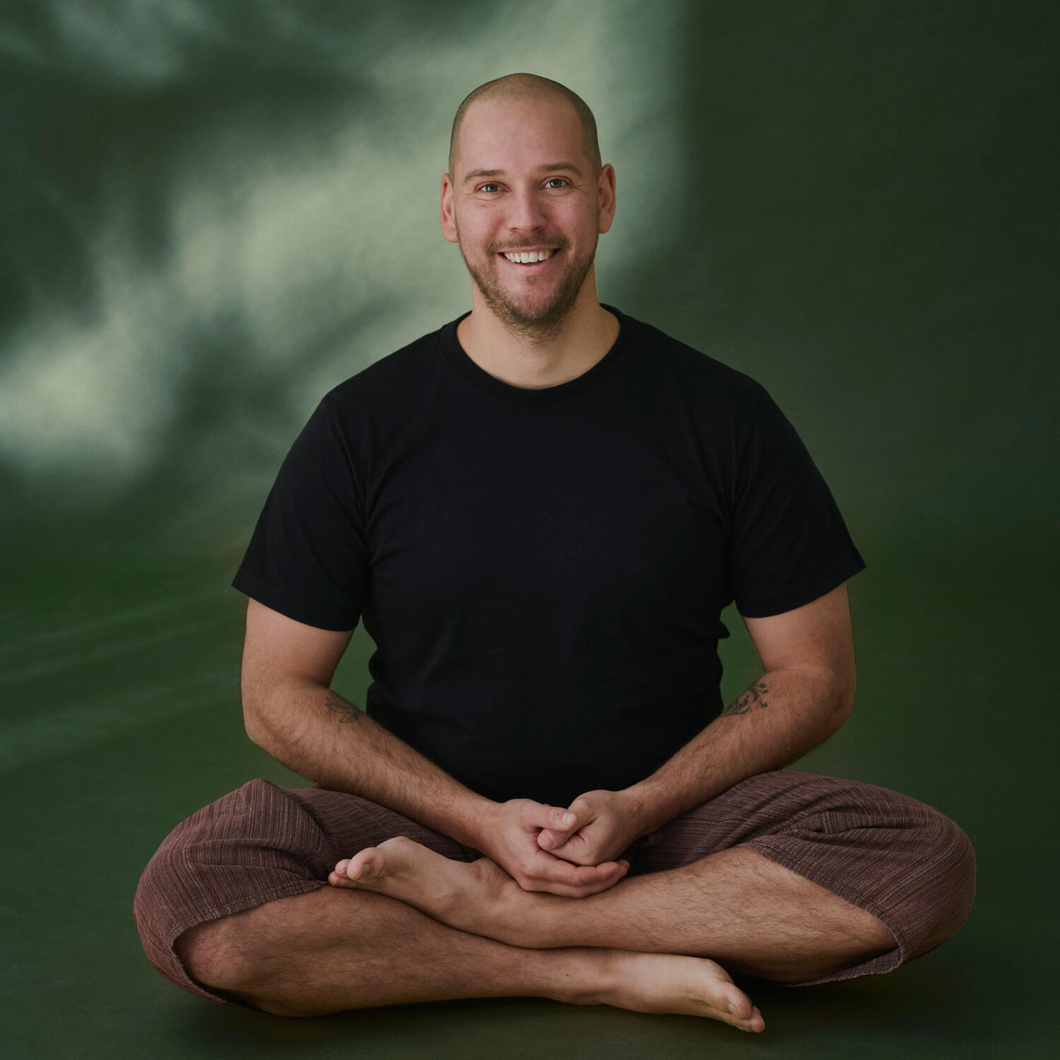 Yoga Kendra Malmö Yoga Teacher, Hatha, Vinyasa, Yin Yoga A smiling, barefoot man sitting cross-legged in a black t-shirt and brown pants against a green background