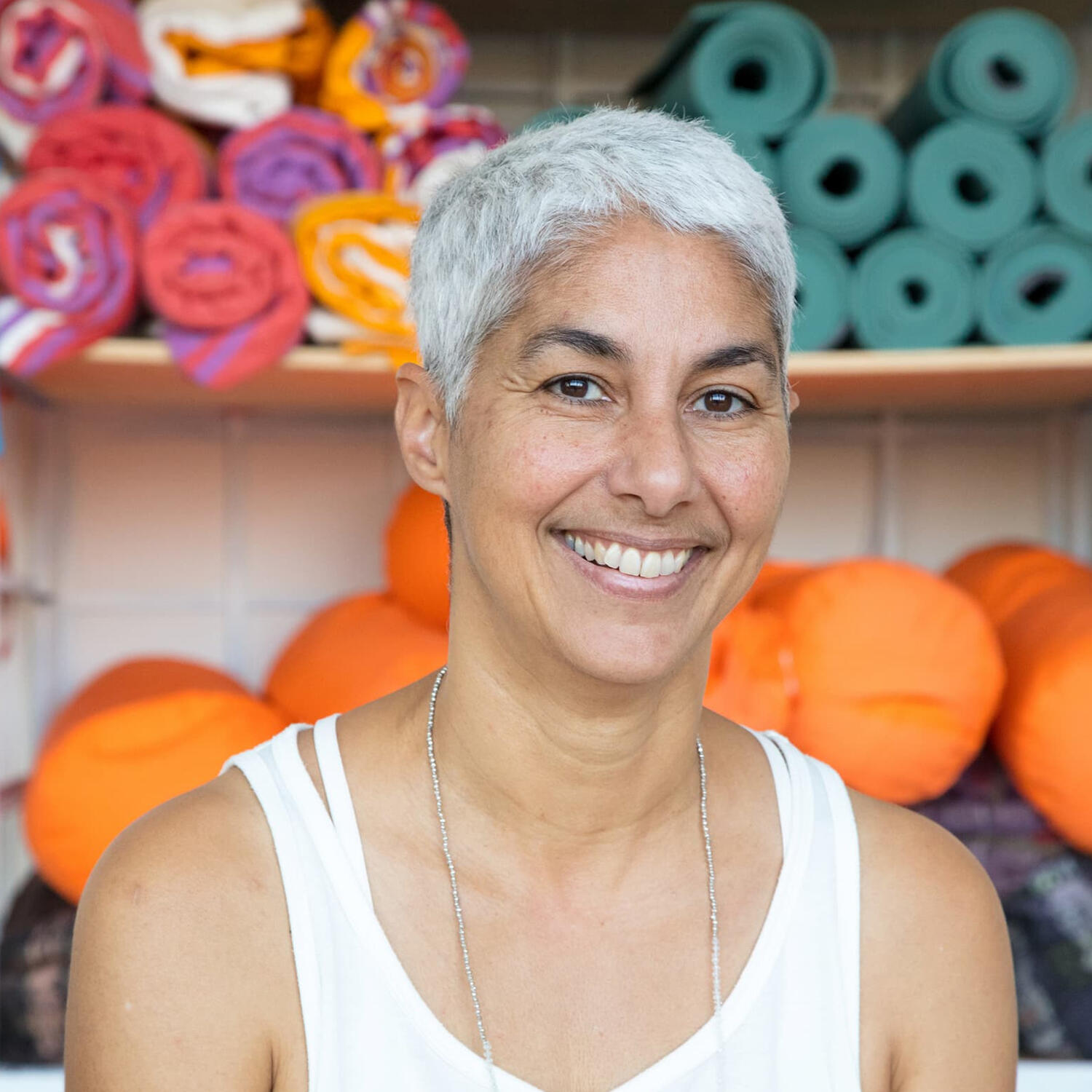 Yoga Kendra Malmö Hatha, Yin, Deep Stretch smiling woman with short grey hair standing in front of shelves filled with yoga mats and props.