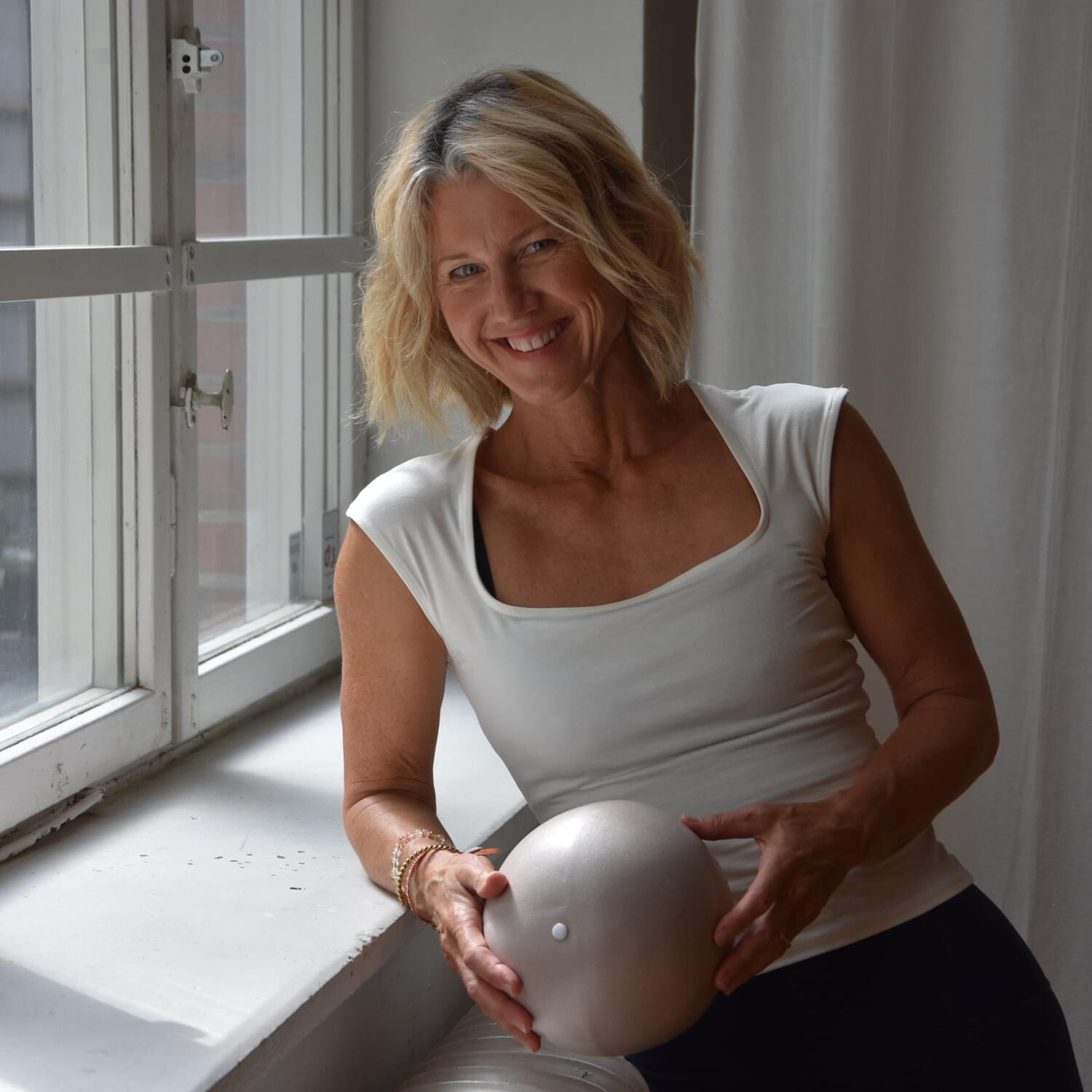 Yoga Kendra Malmö Vinyasa, Barre smiling blonde woman sitting by a window while holding a small, silver-colored exercise ball.
