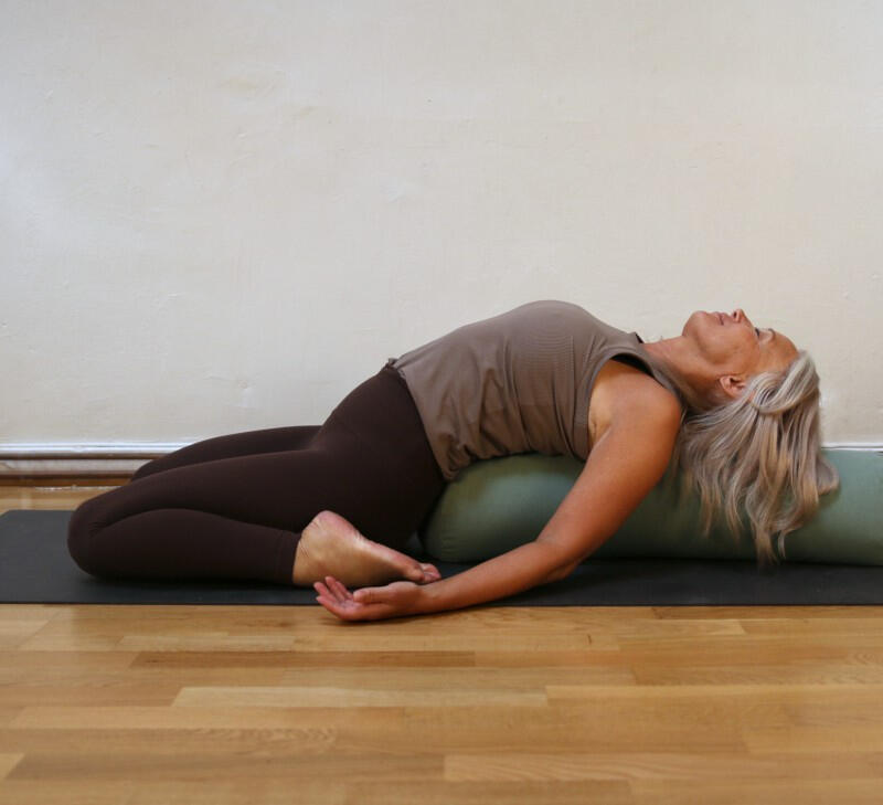 Yin Yoga Kendra Malmö Woman practicing a gentle reclined backbend, supported by using an olive green cylindrical bolster