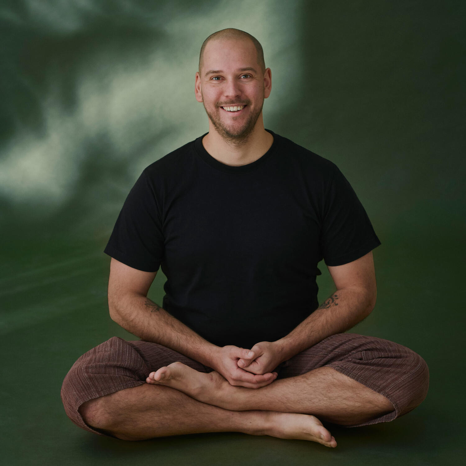 Yoga Kendra Malmö Yoga Teacher, Massage Therapist, Nature Guide A smiling, barefoot man sitting cross-legged in a black t-shirt and brown pants against a green background
